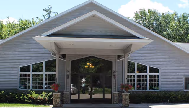 Front exterior view of a single-story building with a peaked roof and large glass entrance doors. The building has light gray siding, multiple windows, stone pillars supporting a covered entryway, and landscaping with bushes and flowers. Trees and a partly cloudy sky are visible in the background.
