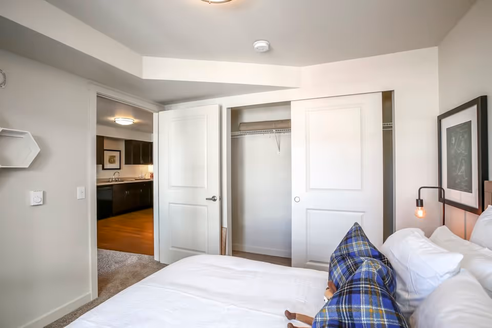 A bright bedroom with a white bed and blue plaid pillows. The room features a closet with sliding doors, one door open revealing a shelf and hanging rod. The bedroom door is open, showing a view into a kitchen area with dark cabinets and wooden flooring. A framed picture and a wall-mounted lamp are visible beside the bed.