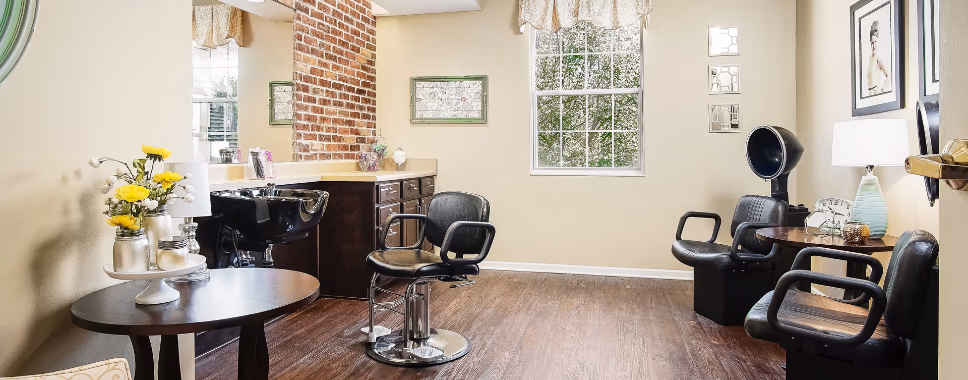 A bright and clean hair salon area inside a senior living facility featuring a black salon chair in front of a black hair washing sink, a small round table with yellow flowers in vases, and additional black chairs with a hair dryer hood. The room has light beige walls, a window with a valance, framed pictures, and wood flooring.