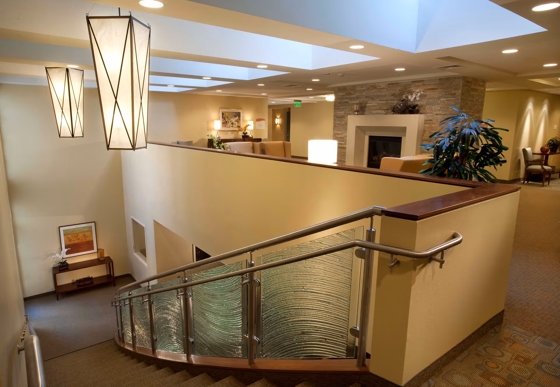 Interior view of a senior living facility showing a staircase with a glass and metal railing, modern hanging pendant lights, a seating area with armchairs near a stone fireplace, and decorative plants and artwork on the walls.