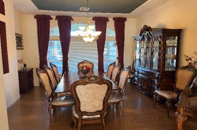 A formal dining room with a long wooden dining table surrounded by eight upholstered chairs. The room features large windows with closed blinds and burgundy curtains. A chandelier hangs above the table, and a wooden china cabinet filled with glassware is against the right wall. The floor is wooden, and there are decorative elements such as a flower arrangement on the table and framed artwork on the walls.