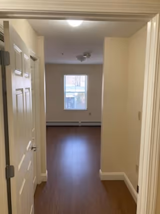 View through an open doorway into an empty room with wood flooring, beige walls, a window at the far end, and ceiling light fixtures.