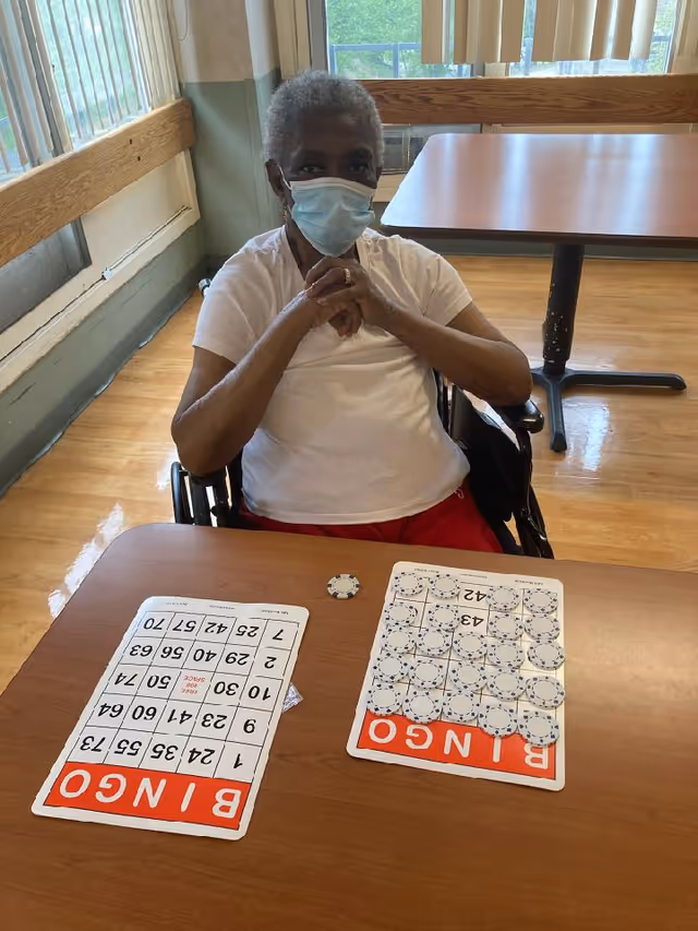 An elderly person wearing a face mask sits in a wheelchair at a table with two bingo cards and a bingo chip in front of them in a well-lit room with large windows and wooden floors.
