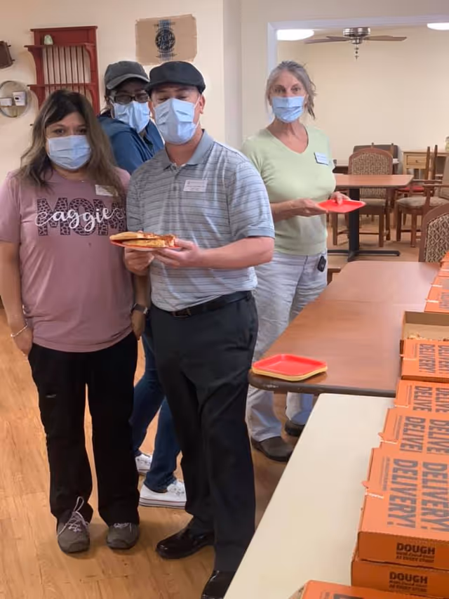 Four people wearing face masks standing indoors near a table with multiple pizza boxes. One person is holding a plate with pizza slices. The room has wooden flooring and dining tables with chairs in the background.