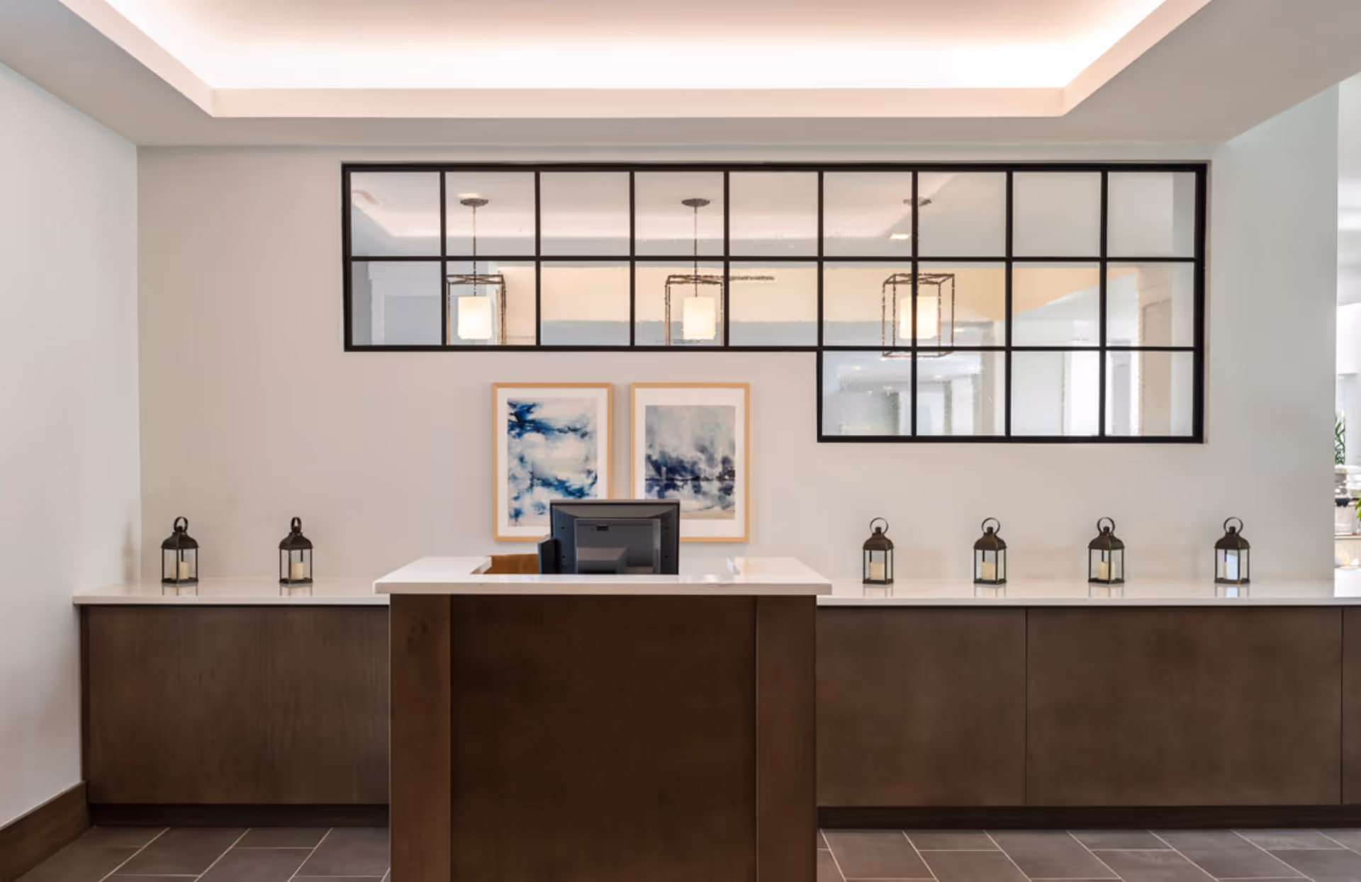 Reception desk in a modern senior living lobby with lanterns, framed artwork, and a large interior window above.