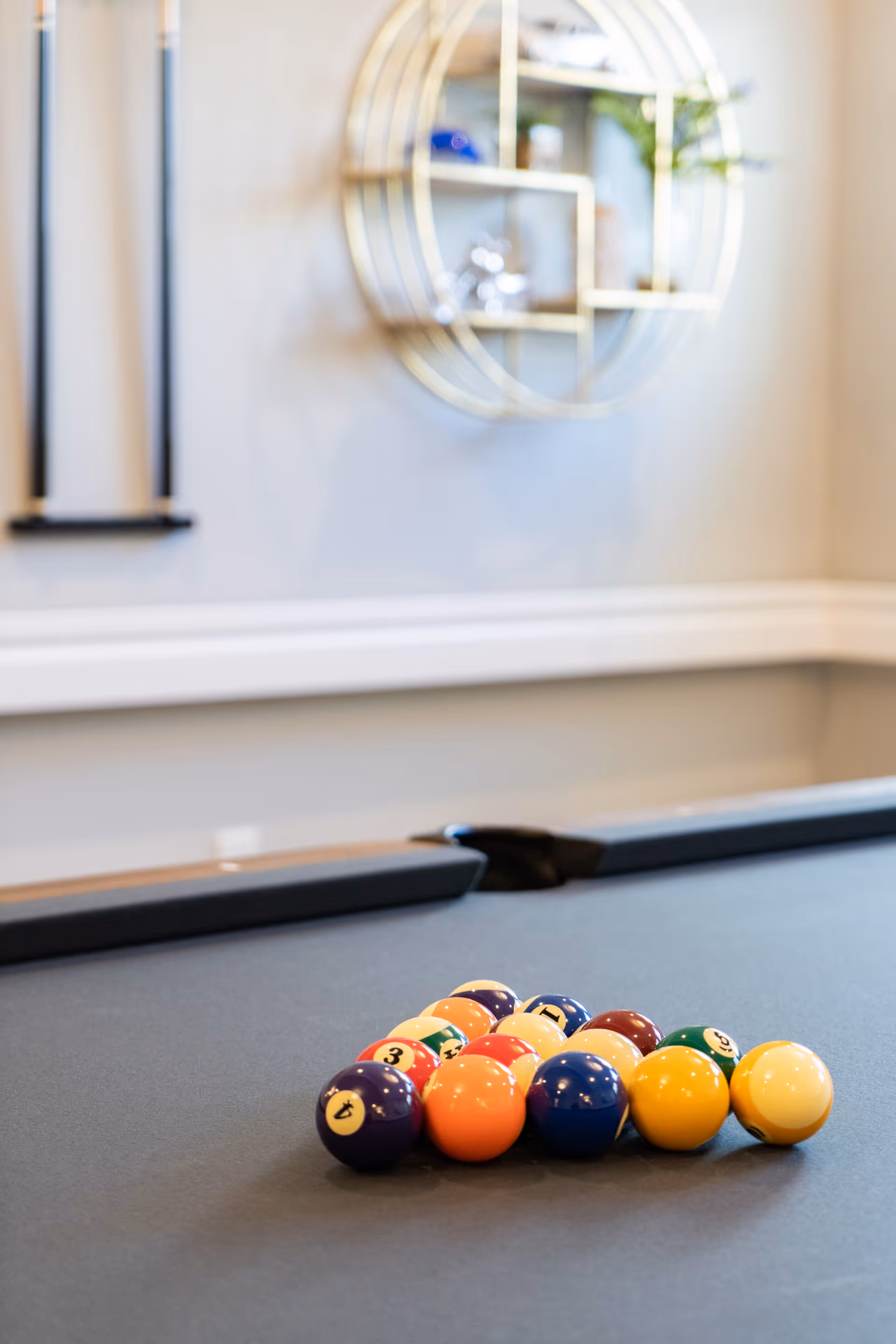 A close-up view of a pool table with a neatly racked set of colorful billiard balls ready for a game. In the background, there is a decorative circular wall shelf and a wall-mounted cue rack holding pool cues.