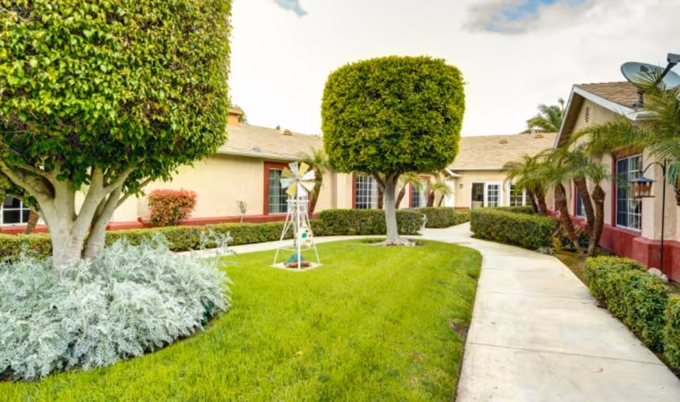 A well-maintained outdoor garden area at Ivy Terrace at Santa Ana Memory Care featuring neatly trimmed trees, green grass, a small decorative windmill, and a curved concrete walkway surrounded by bushes and palm trees, with beige buildings in the background.