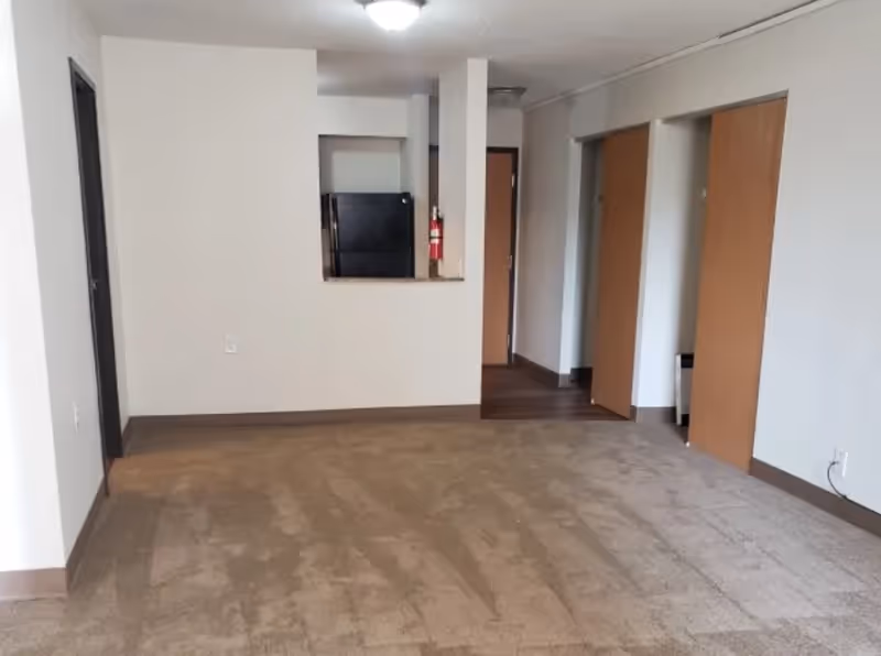 Empty living room with beige carpet, a pass-through to a kitchen showing a black refrigerator, and two open closet doors.