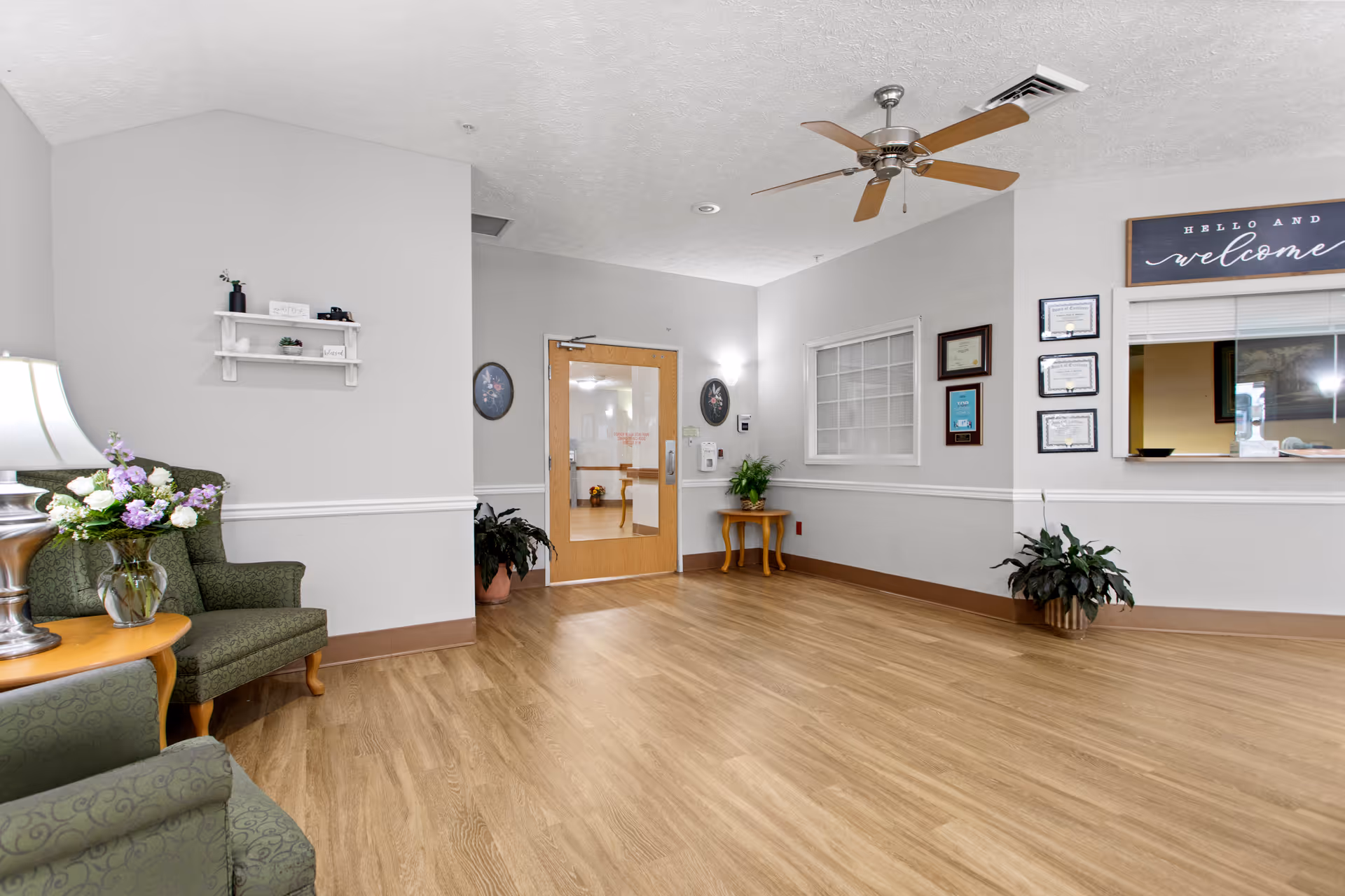 A clean and well-lit waiting area with light wood flooring, two green upholstered armchairs, a small wooden side table with a lamp and a vase of flowers, a ceiling fan, and a reception window with a 'Hello and welcome' sign above it. There are framed certificates on the wall and a wooden door with glass panels leading to another room.