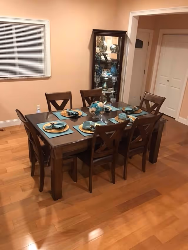 A dining room with a wooden table set for six people. Each place setting has a gold charger plate, a teal placemat, and a teal napkin with a napkin ring. There is a centerpiece with decorative items on the table. The room has hardwood floors, peach-colored walls, a window with blinds, and a glass display cabinet in the corner.