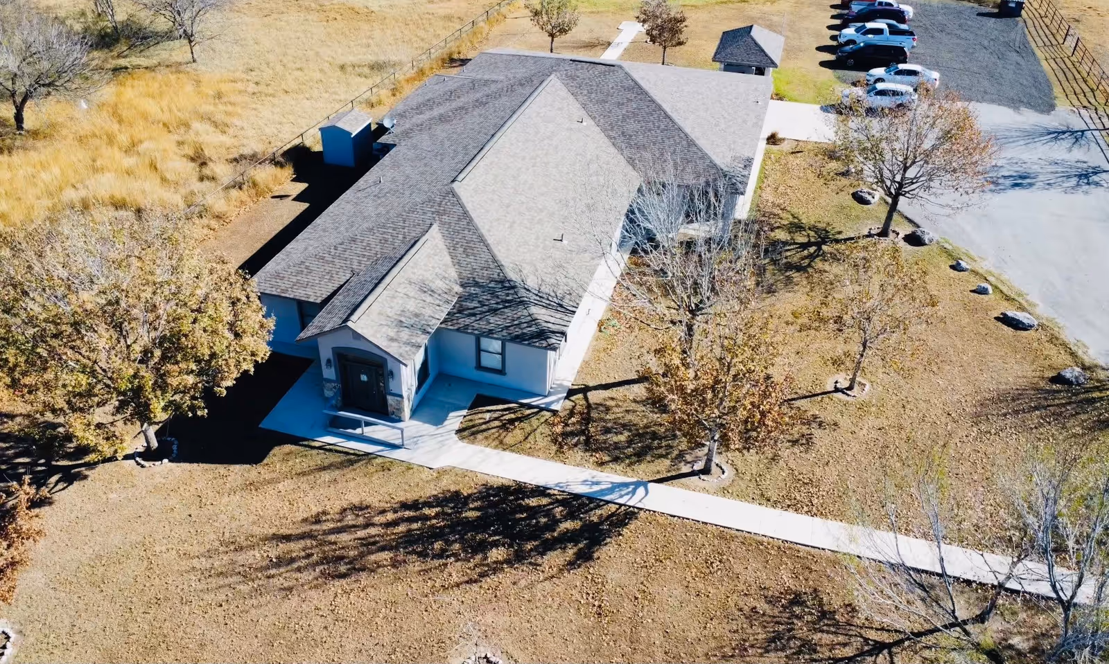 Aerial view of a single-story building with a gray roof surrounded by a grassy area with several trees and a paved walkway leading to the entrance. There is a small parking area with several cars parked near the building.