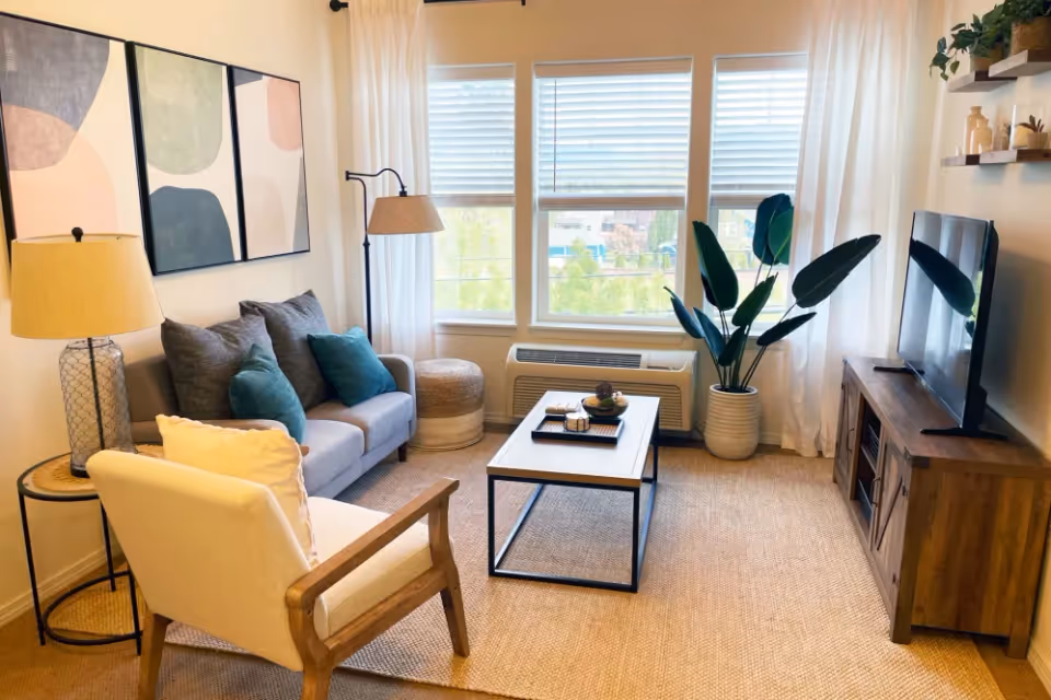 Bright furnished living room with a sofa, armchair, coffee table, TV on a wooden stand, and large windows with blinds.