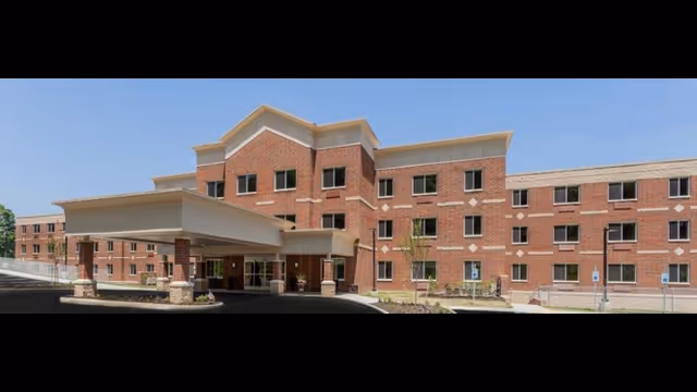 Front exterior of a three-story brick senior living building with a covered porte-cochère entrance under a clear blue sky.