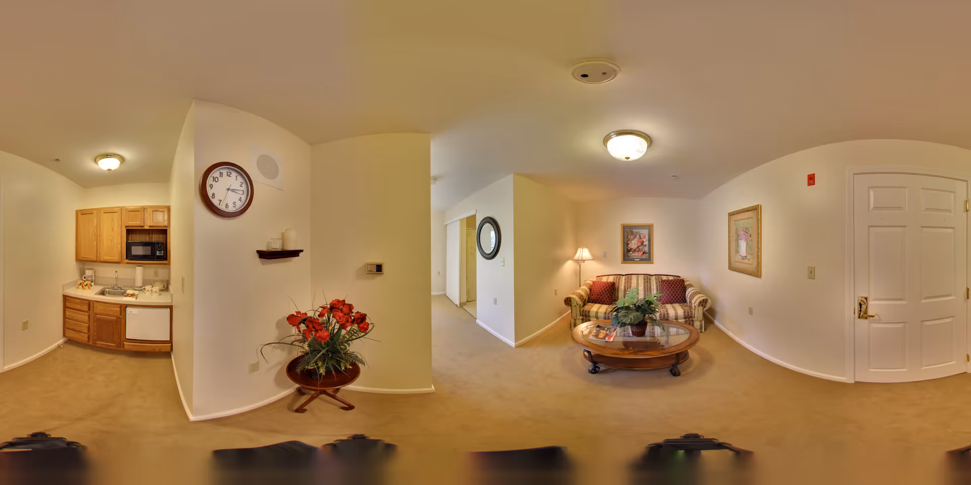 A cozy interior space featuring a small kitchen area with wooden cabinets, a microwave, and a dishwasher on the left. In the center, there is a wall with a round clock and a small shelf holding candles above a table with a red floral arrangement. To the right, a living room area includes a striped sofa, a round wooden coffee table with a plant, a floor lamp, and framed artwork on the walls. The space is carpeted and well-lit with ceiling lights.