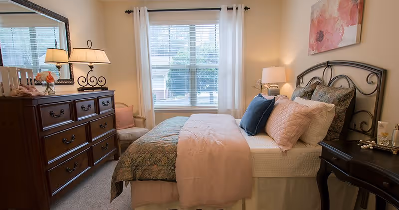 A cozy bedroom with a neatly made bed featuring multiple pillows and a pink throw blanket. There is a wooden dresser with a lamp and flowers on top, a large mirror above the dresser, a window with white curtains letting in natural light, and a small side table with a lamp and decorative items.