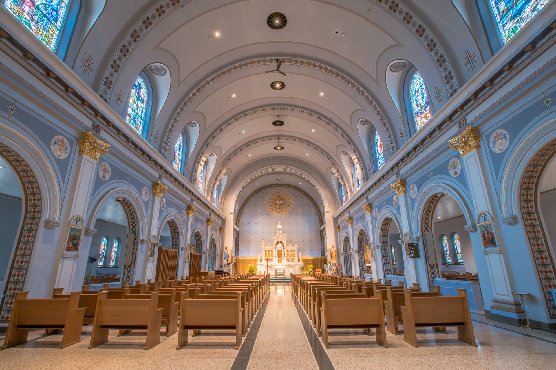 Interior view down the central aisle of an ornate chapel or church with rows of wooden pews, arched ceilings, and stained-glass windows.