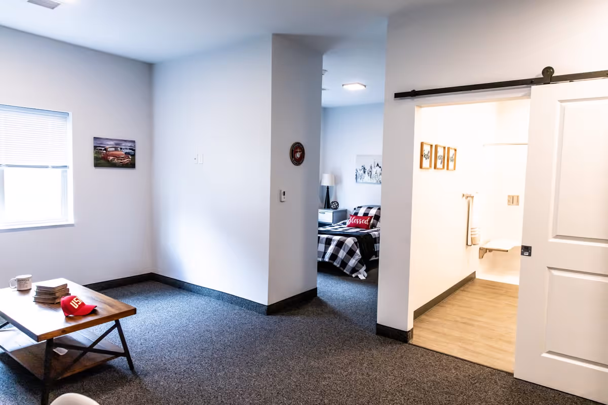 Interior view of a senior living facility room with a small wooden coffee table holding a red cap, books, and a mug. The room has gray carpet and white walls, with a window on the left side. In the background, there is a bedroom with a bed covered in a black and white checkered blanket and a red pillow that says 'blessed'. To the right, there is a doorway leading to a bathroom with light wood flooring and white walls.