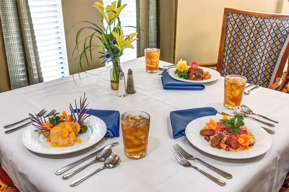 A dining table set for three with white tablecloth, blue napkins, and plates of gourmet food. Each place setting has a glass of iced tea, silverware, and a plate with colorful, artfully arranged dishes. A small vase with yellow lilies and greenery, along with salt and pepper shakers, is placed in the center of the table. The background shows a window with blinds and patterned chairs.