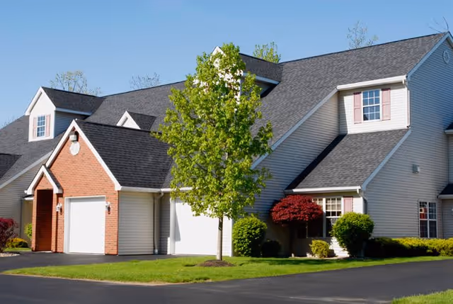 Exterior view of a residential-style building with white siding and brick accents, featuring multiple gabled roofs and dormer windows. There is a tree and some shrubs in front of the building, with a paved driveway and green lawn surrounding it.