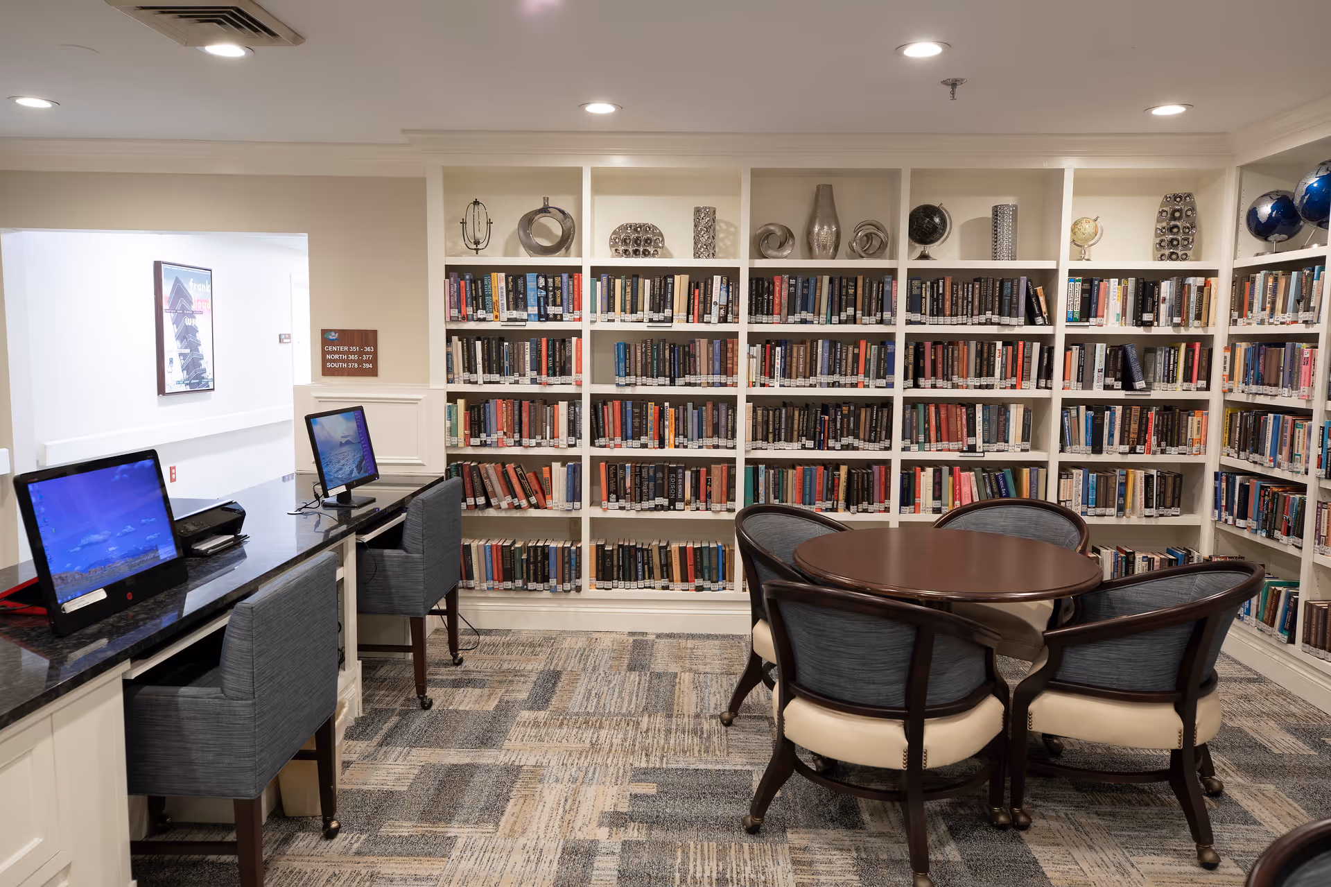A cozy library room in a retirement community featuring white built-in bookshelves filled with books and decorative items. In the center, there is a round wooden table surrounded by four cushioned chairs. On the left side, there is a long desk with two computer monitors and two chairs. The room has a patterned carpet and recessed ceiling lights.