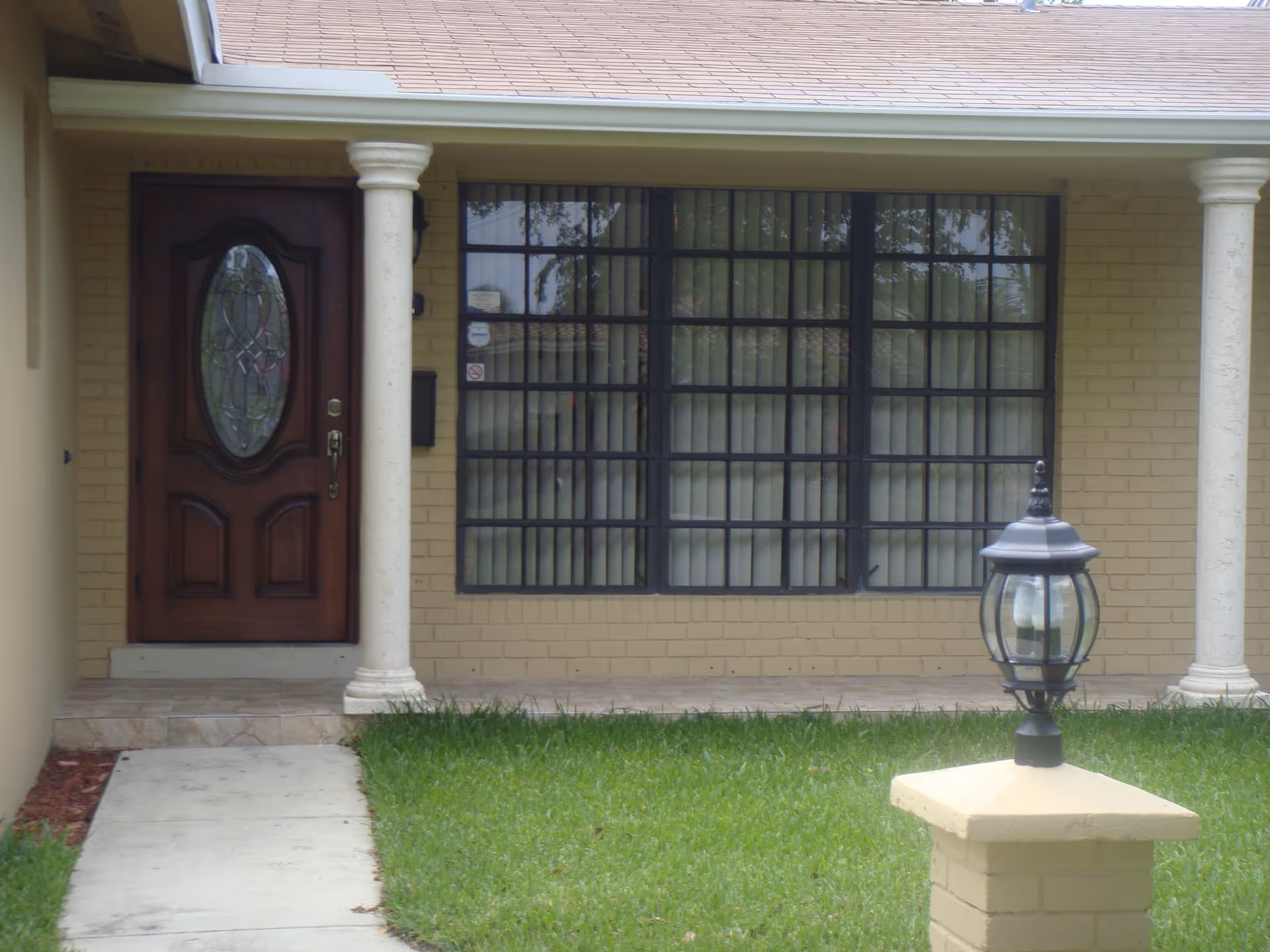 Front entrance of a building with a wooden door featuring an oval glass design, two white columns on either side, a large window with vertical blinds, a tiled porch, green grass lawn, and a black outdoor lamp mounted on a yellow brick post.