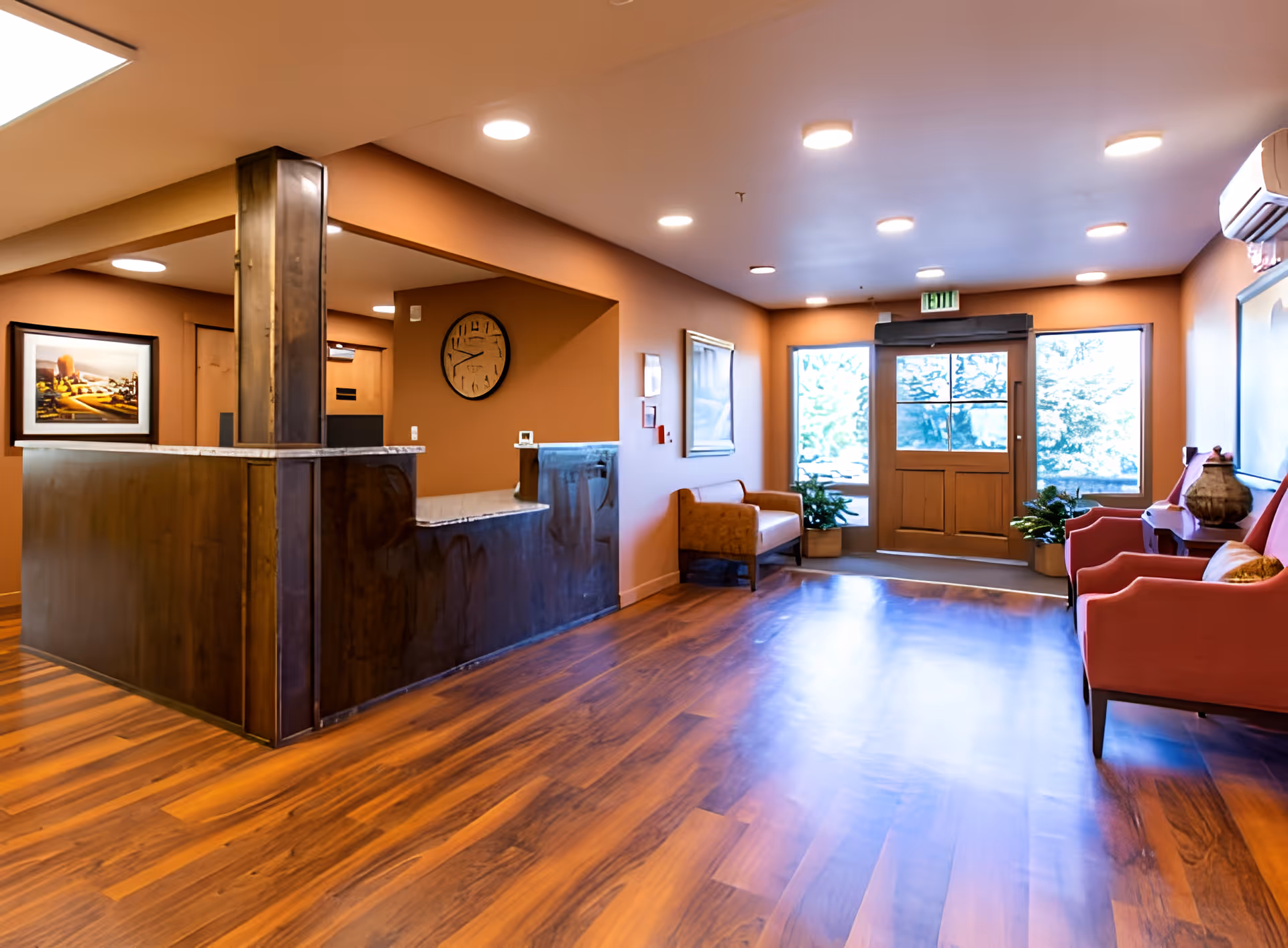 Warm, wood-floored reception lobby with a dark wooden front desk, seating area, and double doors letting in daylight.