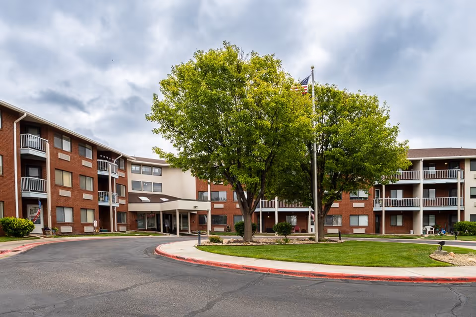 Exterior view of a multi-story senior living facility with red brick and beige walls, balconies, and a circular driveway. There are two large green trees and a flagpole with an American flag in the center of a landscaped grassy area. The sky is cloudy.