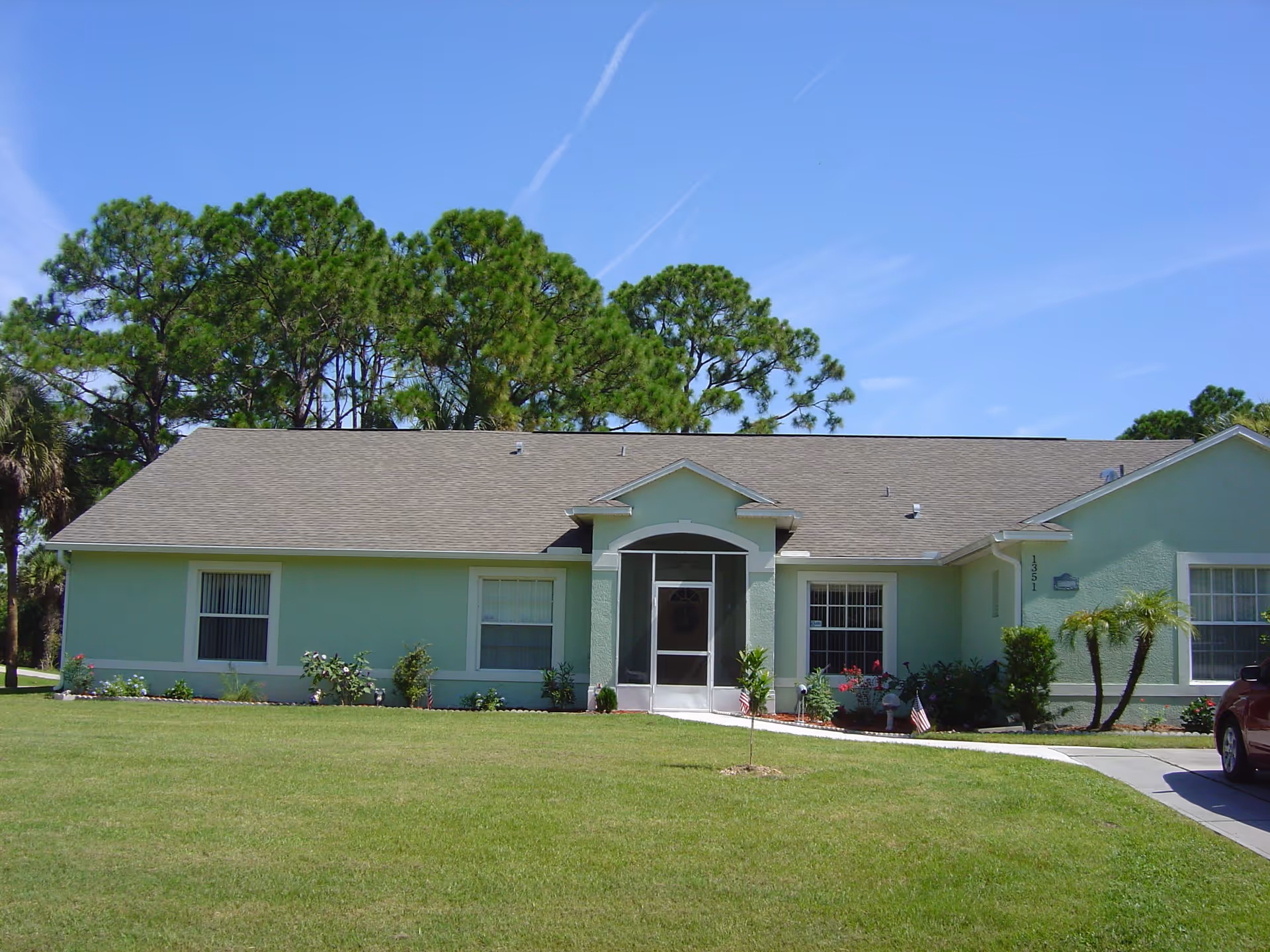 Single-story light green building with a gray shingled roof, a screened front porch entrance, several windows, and a well-maintained lawn with small plants and trees in front. Tall pine trees are visible in the background under a clear blue sky.