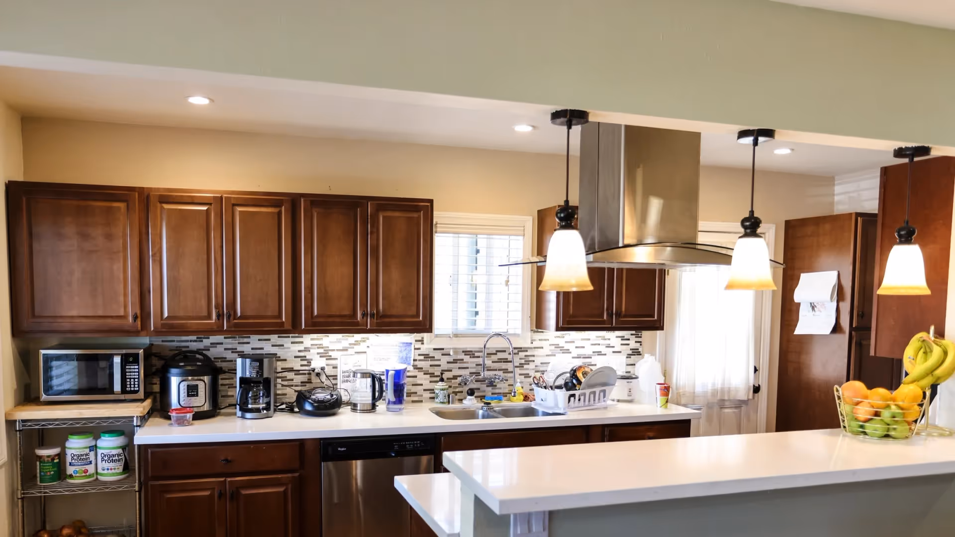 Open kitchen with dark wood cabinets, white countertops, a stainless range hood, pendant lights, and small appliances on the counter.
