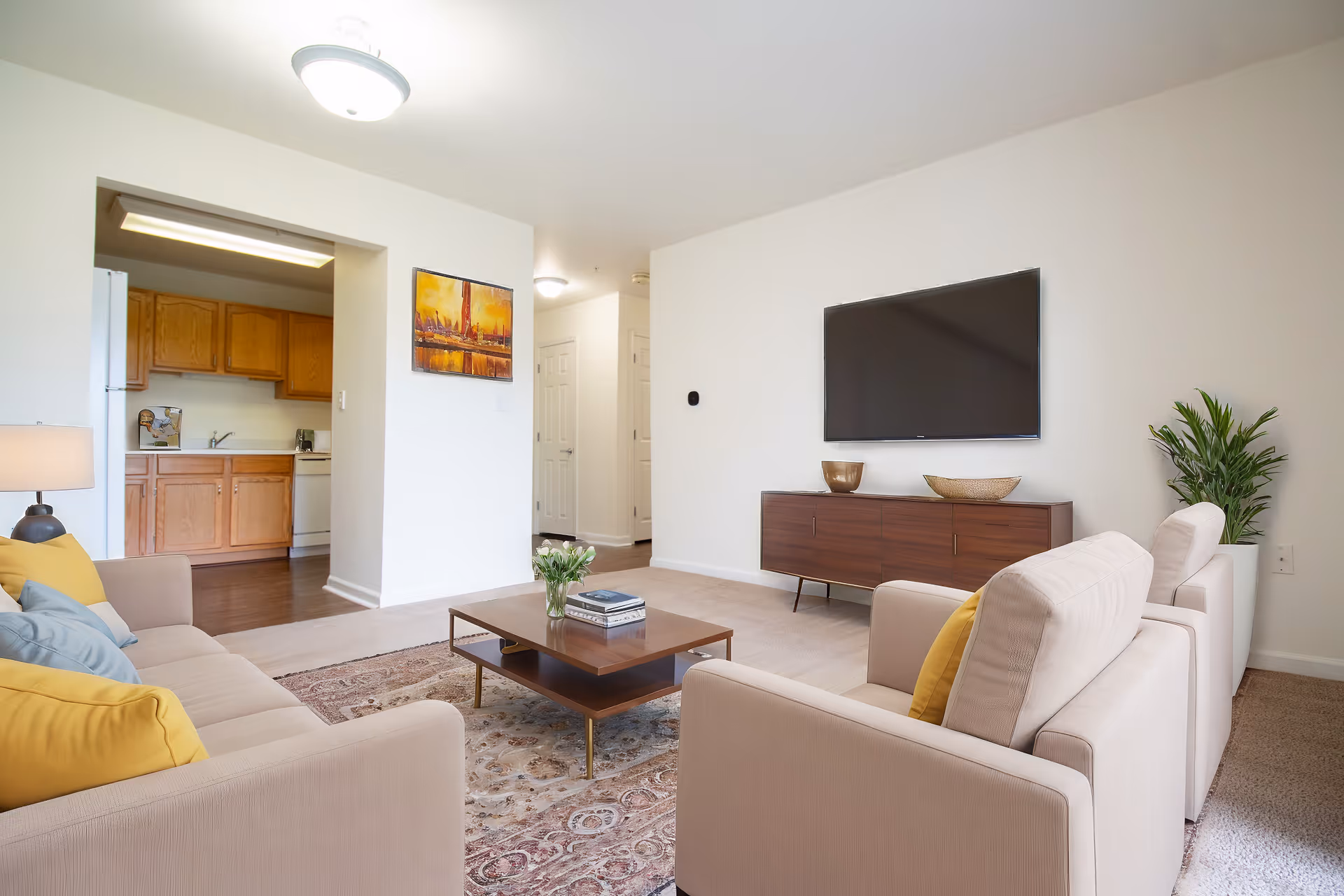 Bright living room with beige sofas, a coffee table, a wall-mounted TV, and a view into the kitchen.
