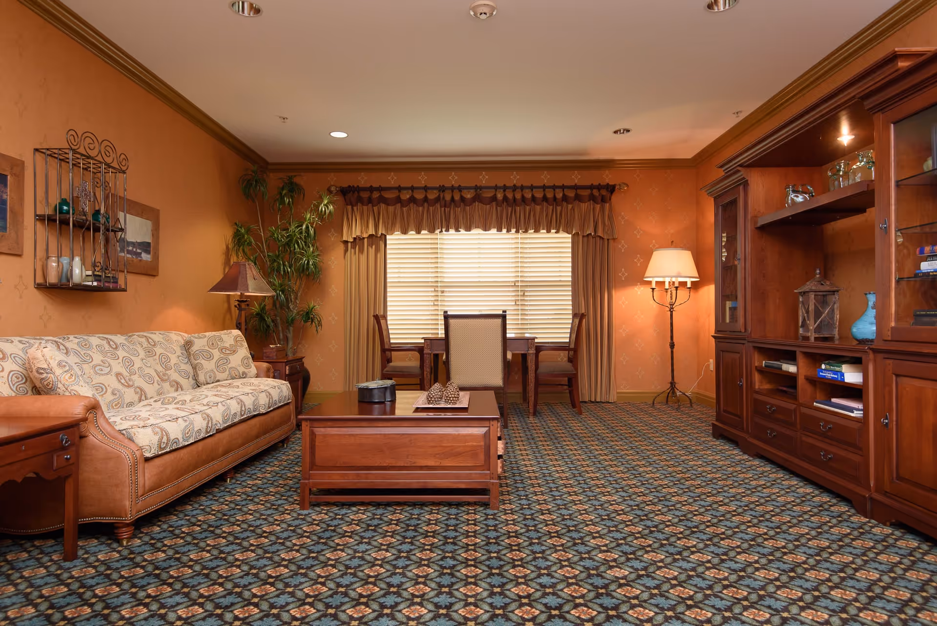 A cozy living room with a patterned carpet, a beige and brown sofa, a wooden coffee table, a wooden entertainment center with shelves and cabinets, a floor lamp, and a small dining table with three chairs near a window with closed blinds and brown curtains.