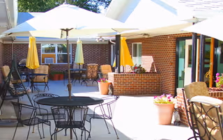 Outdoor patio area with multiple tables and chairs under large white and yellow umbrellas. The space is surrounded by brick walls and windows, with potted plants adding greenery and flowers.