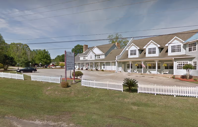 Exterior view of Homestead Village of Pensacola, showing a large two-story building with multiple windows and a white picket fence in front. There is a sign near the entrance displaying the facility's name and contact information. The area includes a parking lot with a few cars and some greenery with trees and grass.