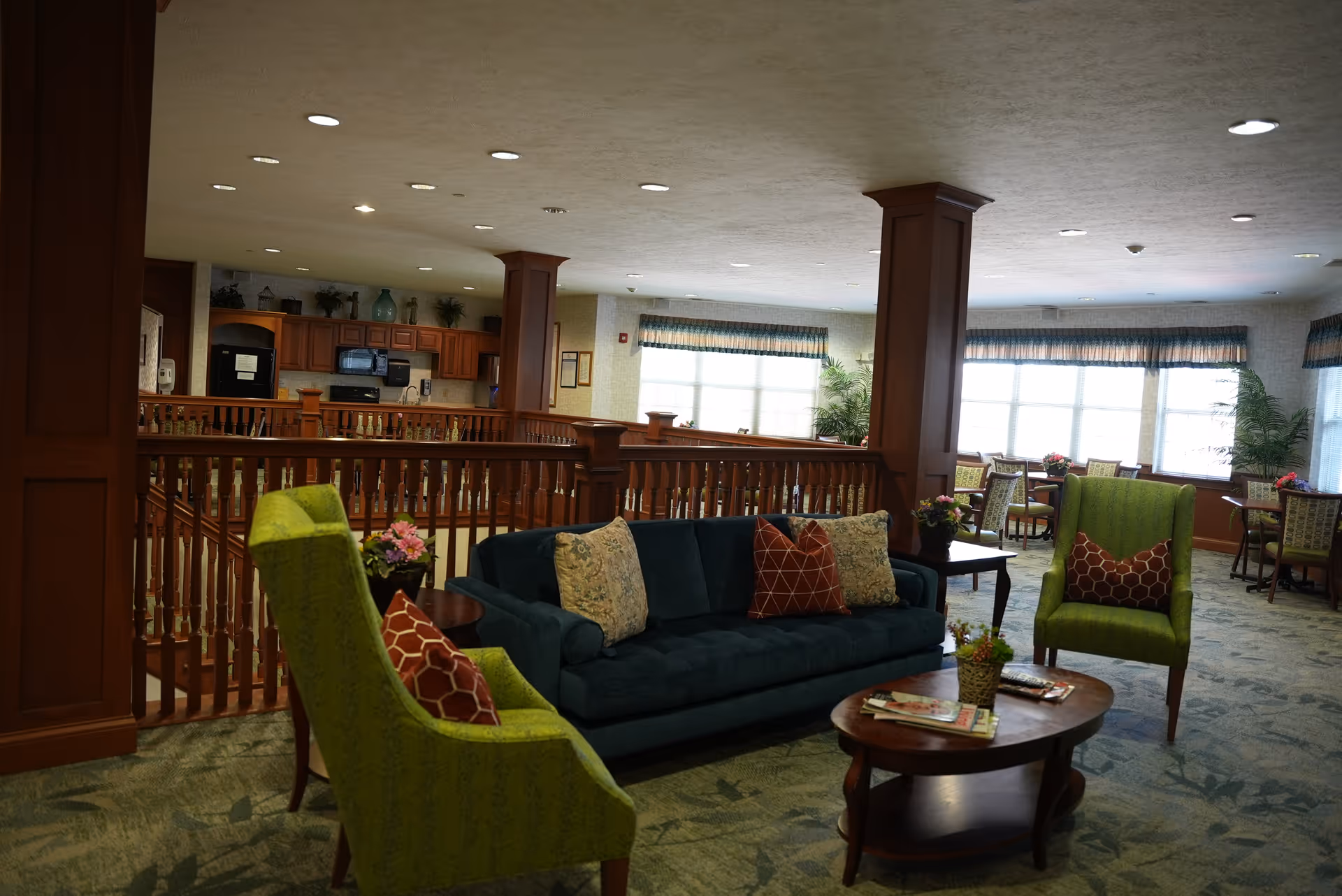A cozy assisted living common area with a dark green sofa and two green armchairs, each with decorative pillows. There is a wooden coffee table with magazines and a small plant on it. The room has large windows with valances, letting in natural light, and a kitchen area with wooden cabinets and appliances in the background. The space is carpeted and features wooden railings and columns.