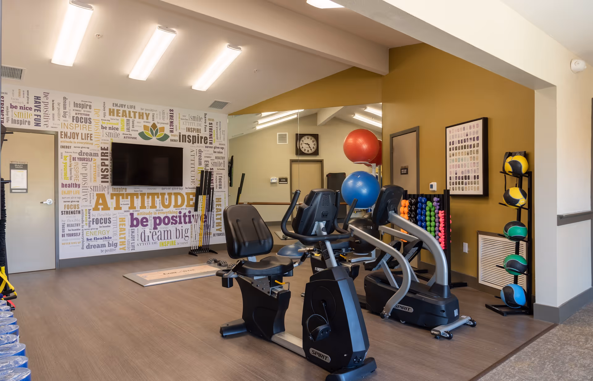 A fitness room in an assisted living facility featuring exercise equipment including stationary bikes, a rack of colorful dumbbells, medicine balls, and large exercise balls. The walls are decorated with motivational words and phrases such as 'attitude', 'be positive', and 'dream big'. A flat-screen TV is mounted on the wall, and the room has bright overhead lighting.