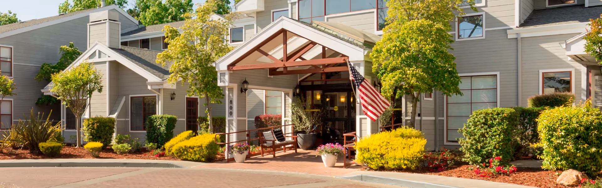 Front entrance of a light-gray senior living building with a covered porte-cochere, American flag, benches, and well-kept landscaping.