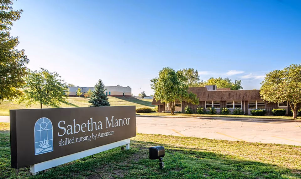 Sign reading 'Sabetha Manor skilled nursing by Americare' in front of the single-story Sabetha Manor building with lawn and trees under a blue sky.