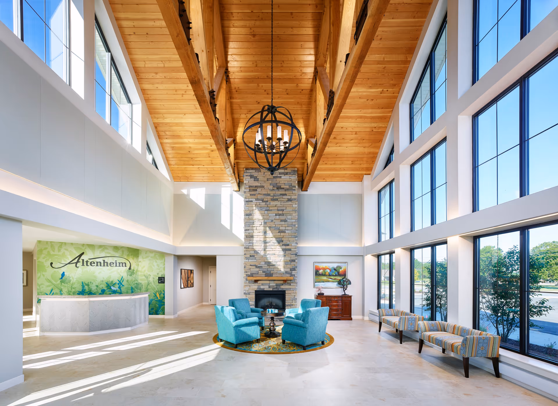 Spacious senior living facility lobby with high wooden ceiling beams, large windows letting in natural light, a stone fireplace, four turquoise armchairs arranged around a small table on a circular rug, and a reception desk with a green floral backdrop displaying the name Altenheim.
