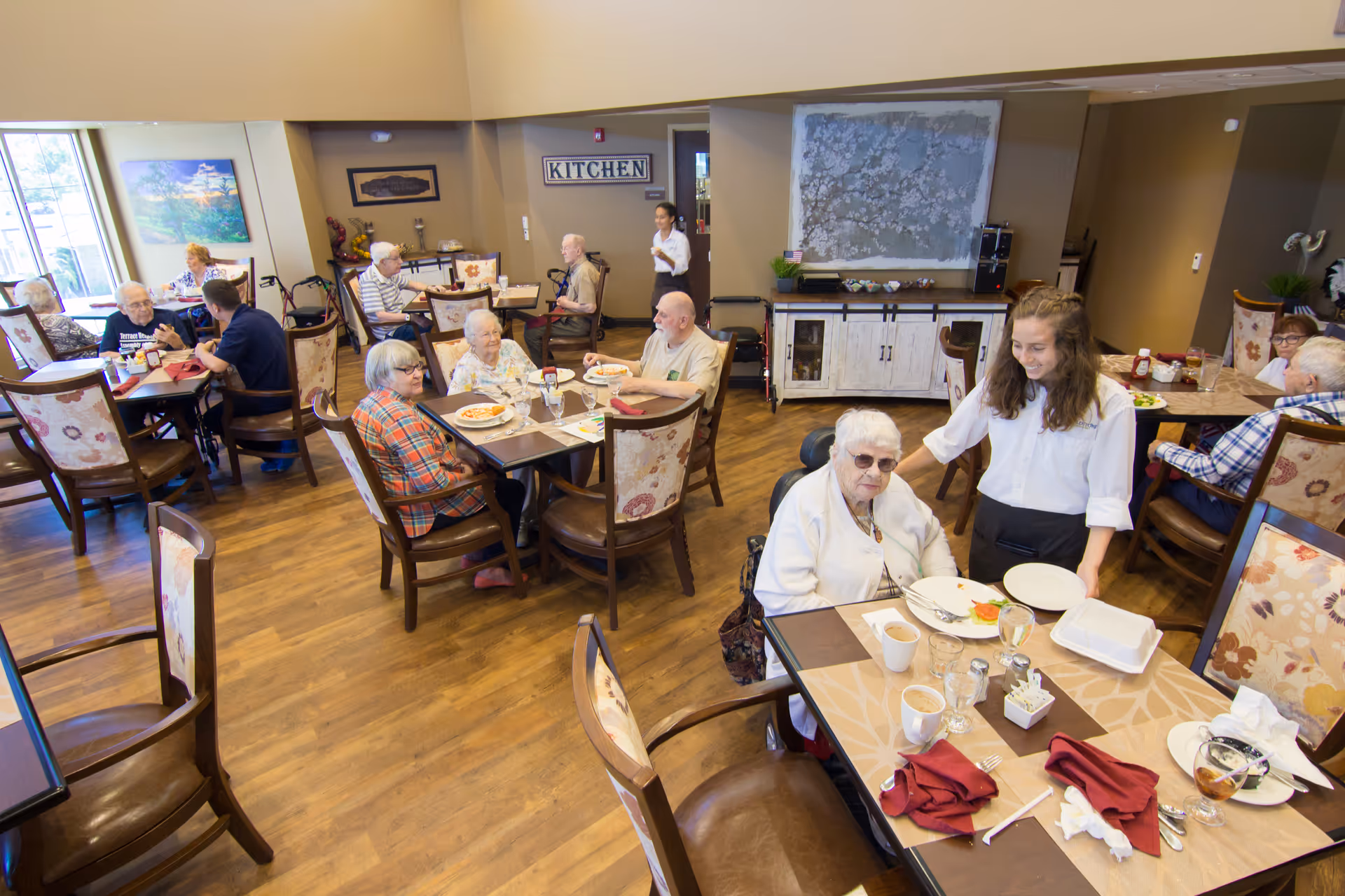 A dining room in a senior living facility with elderly residents seated at tables enjoying meals. A staff member is standing next to one of the residents, smiling. The room has wooden floors, floral upholstered chairs, and a sign on the wall that says 'KITCHEN'.