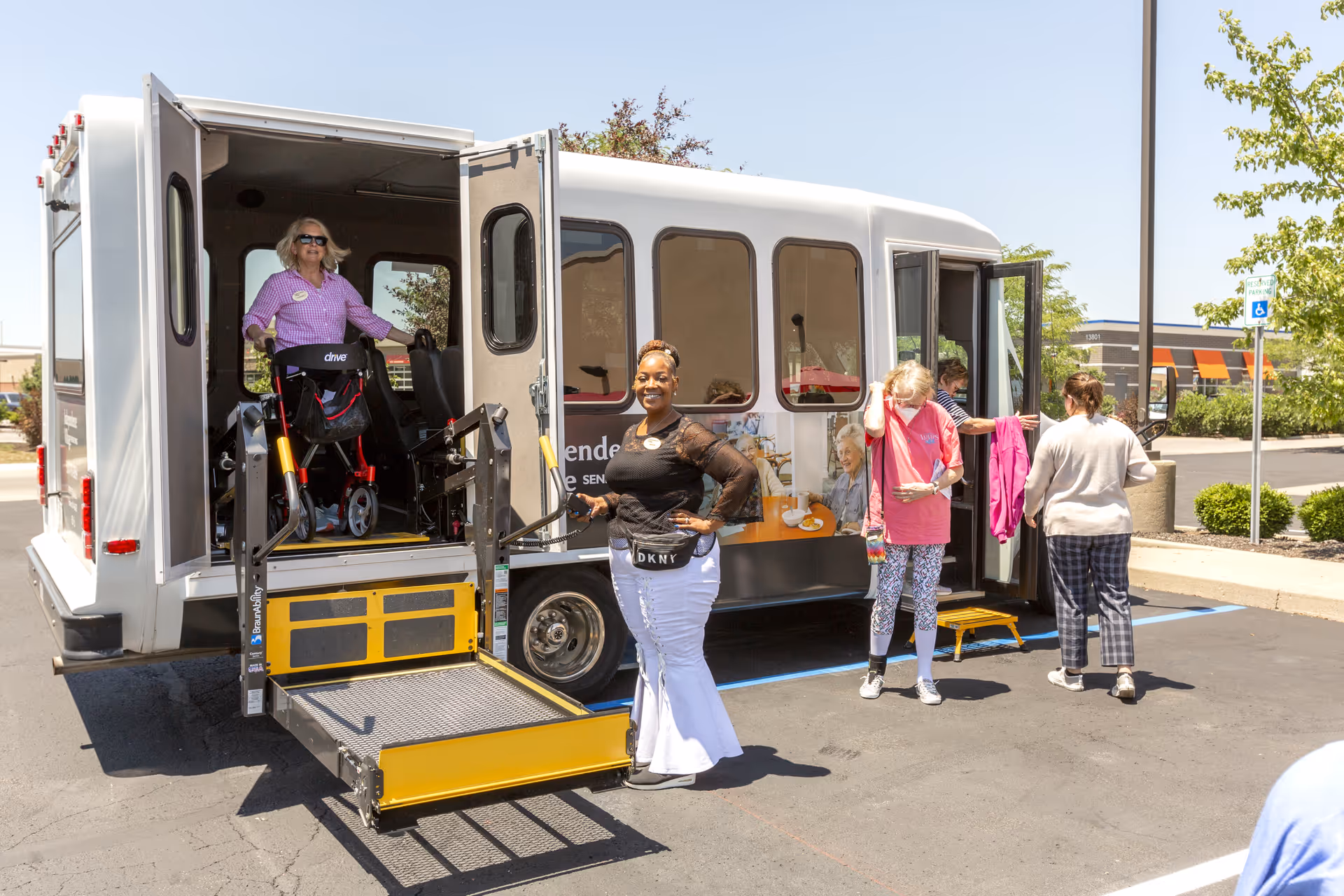 A group of people outside a white shuttle bus with an extended wheelchair lift. One woman is standing on the lift with a walker, another woman stands smiling in front of the lift, and two other women are near the bus entrance. The scene is in a parking lot on a sunny day.