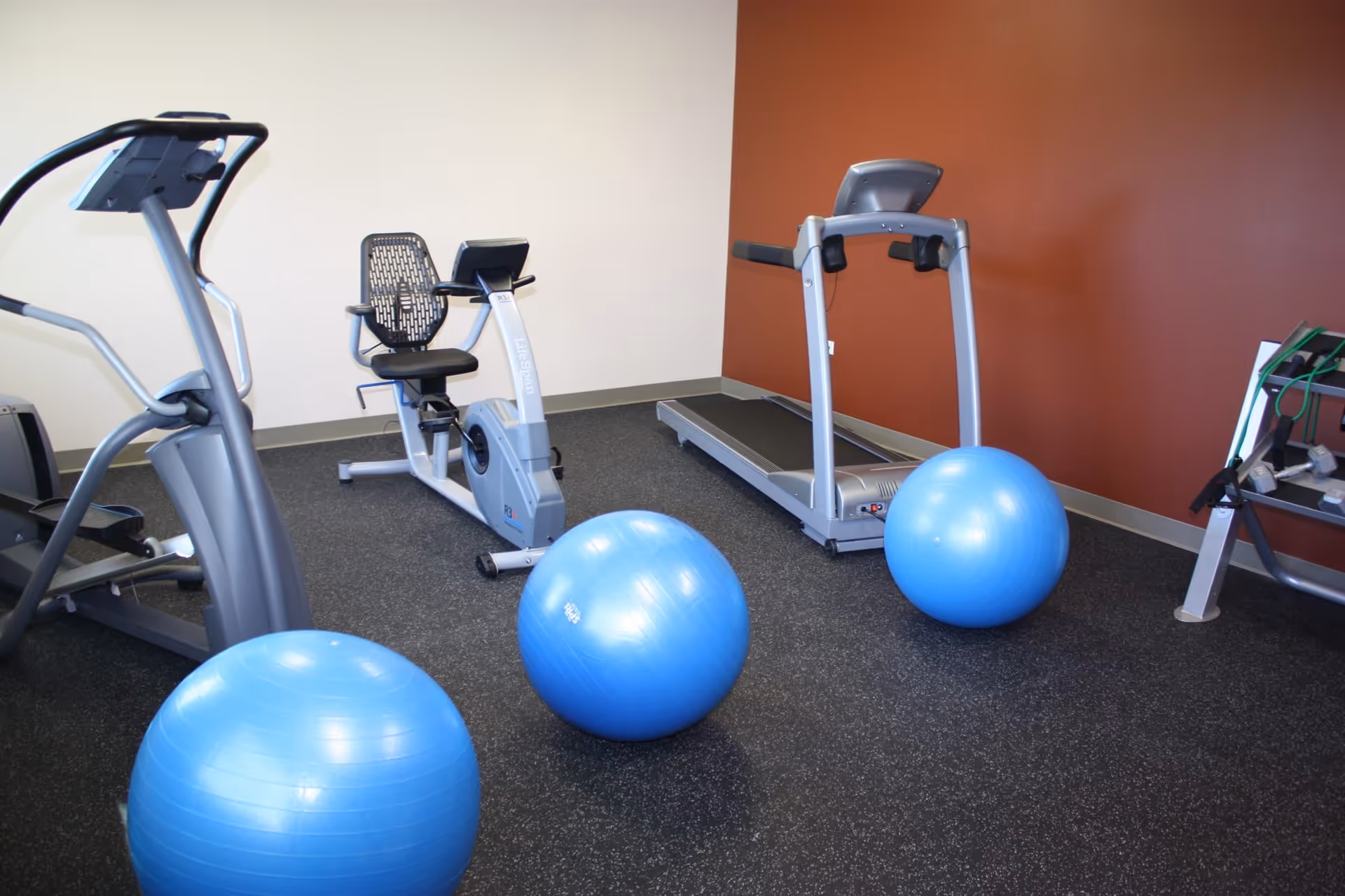 A small fitness room with exercise equipment including a treadmill, a recumbent bike, an elliptical machine, and three blue exercise balls on a black rubber floor. The walls are painted white and orange.