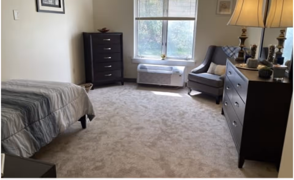 A bedroom with a bed covered in a gray and white striped comforter on the left, a dark wooden chest of drawers against the far wall, a window with blinds letting in natural light, a gray armchair with a pillow and a blanket draped over the back, and a dark wooden dresser with a mirror and decorative items on the right.