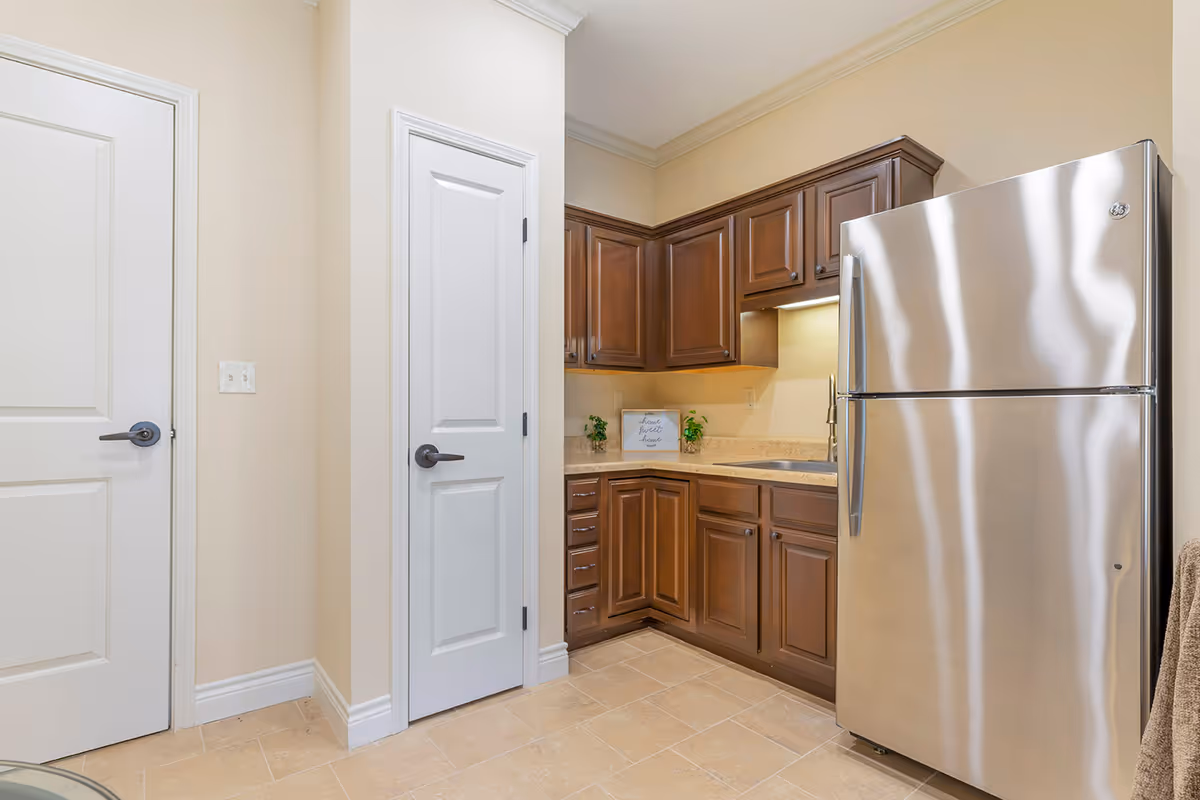 A small kitchen area with dark wooden cabinets, a beige countertop, and a stainless steel refrigerator. There are two white doors on the left side of the image, beige tiled floor, and a small framed sign on the countertop that says 'home sweet home' with two small potted plants beside it.