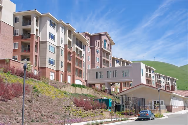 Exterior view of a multi-story retirement community building with a sky bridge connecting two sections. The building is situated on a hillside with landscaped plants and a clear blue sky overhead. A car is parked near the entrance of the building.