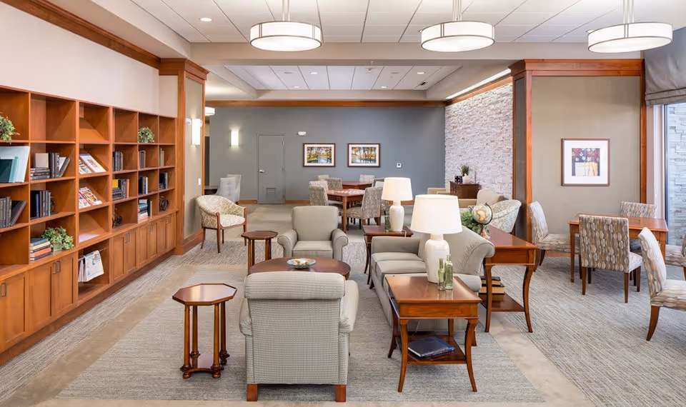 A bright and spacious senior living common area featuring comfortable armchairs and sofas arranged around wooden coffee and side tables with lamps. There is a large wooden bookshelf filled with books and decorative plants on the left wall. In the background, there are additional seating areas with tables and chairs, framed artwork on the walls, and a stone accent wall on the right side near large windows with curtains.