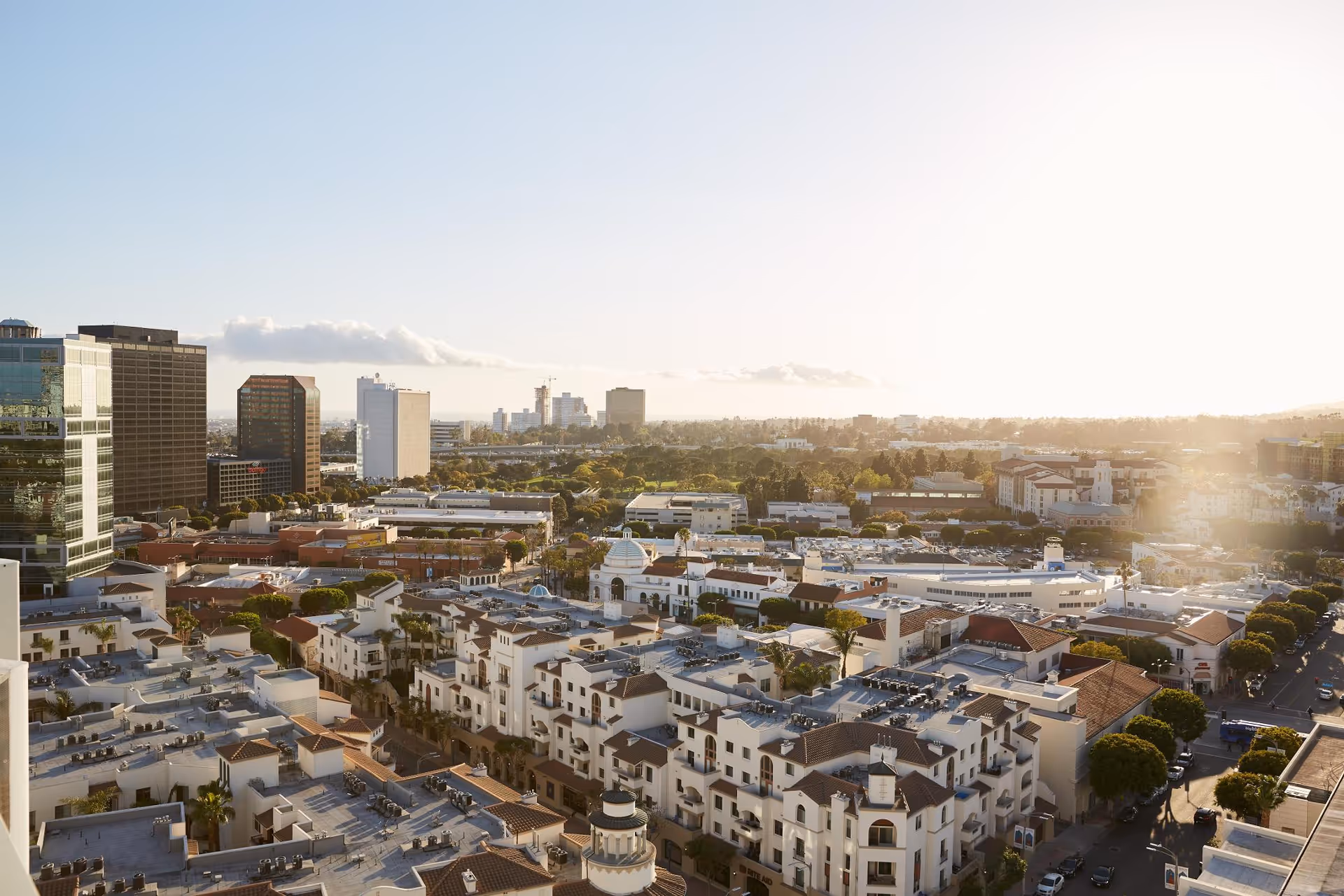 Aerial view of a cityscape featuring a mix of modern high-rise buildings and Mediterranean-style residential buildings under a clear sky with the sun shining brightly from the right side.