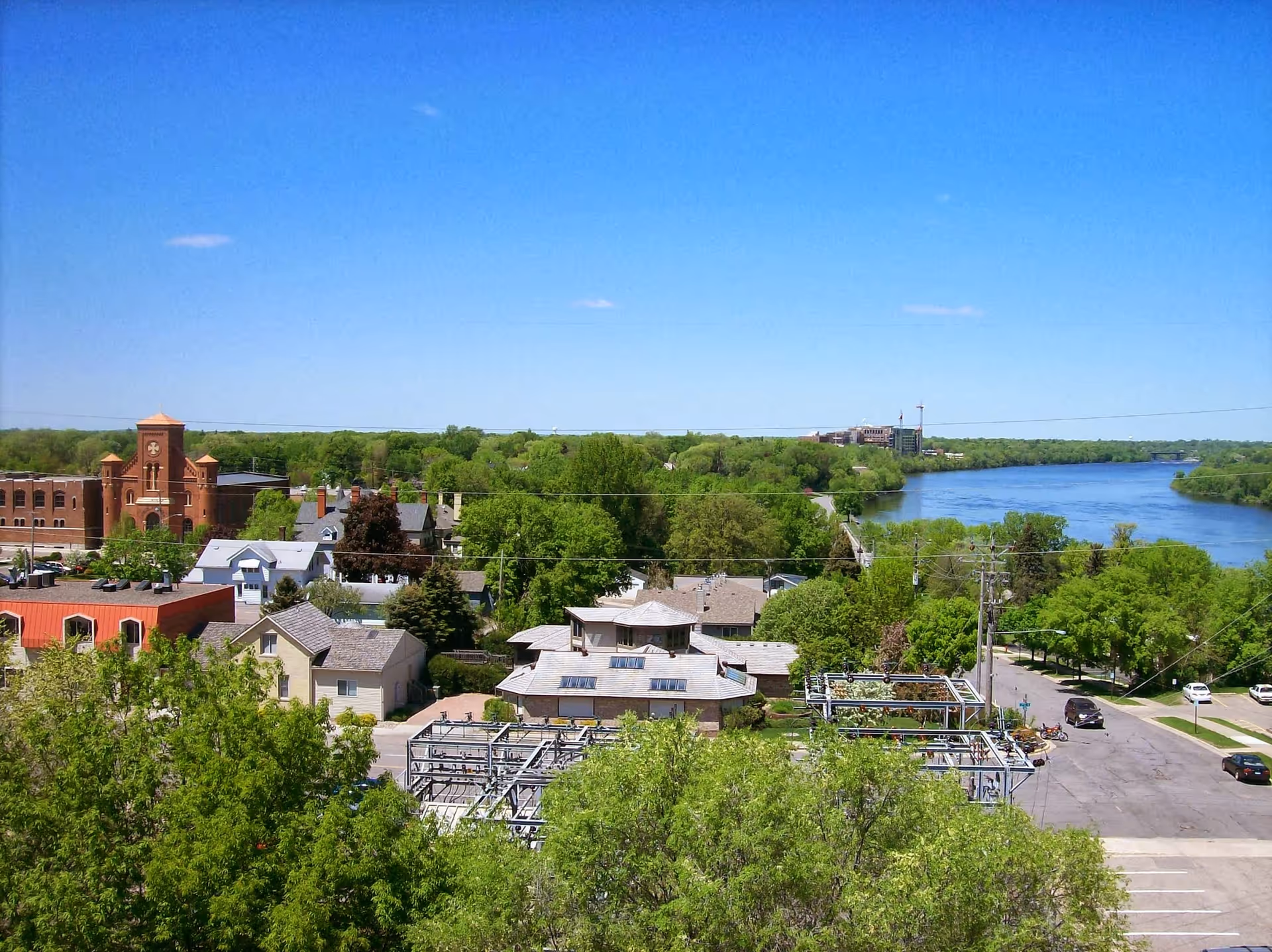 A scenic view of a small town with various buildings, lush green trees, and a river flowing on the right side under a clear blue sky.
