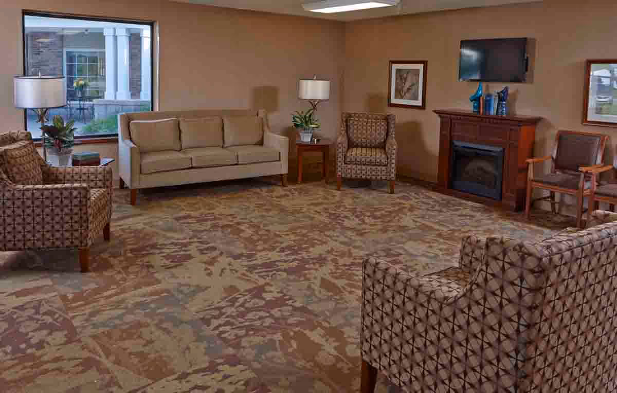 A cozy living room with a beige sofa, patterned armchairs, a wooden fireplace with a TV mounted above it, two framed pictures on the walls, a window showing an outdoor patio, and two table lamps on side tables.