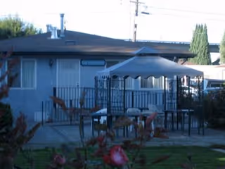 Outdoor patio area at Bethesda Home featuring a canopy-covered seating area with tables and chairs, surrounded by a black metal fence. The building is visible in the background with windows and a door, and some plants and bushes are in the foreground.