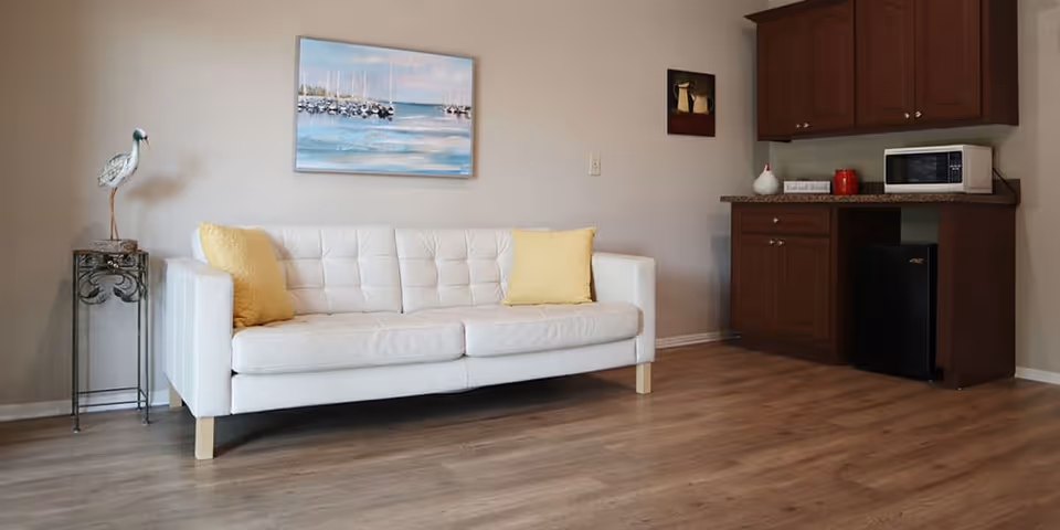 Small living area with a white sofa and yellow pillows, wall art, decorative side table, and a kitchenette with microwave and mini-fridge.
